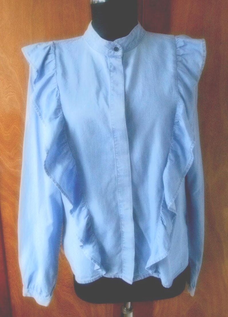 May include: Light blue blouse with a high collar and long sleeves. The blouse features ruffled accents along the shoulders and down the front. The fabric appears to be a lightweight material, suitable for warmer weather.