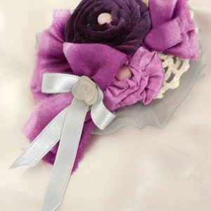 May include: A purple and silver fabric flower brooch with a white pearl center. The brooch is made of layers of fabric and lace.