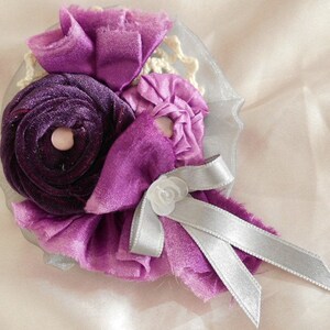 May include: A purple and silver fabric flower brooch with a white center. The flower is made of multiple layers of fabric and has a silver ribbon tied around it.