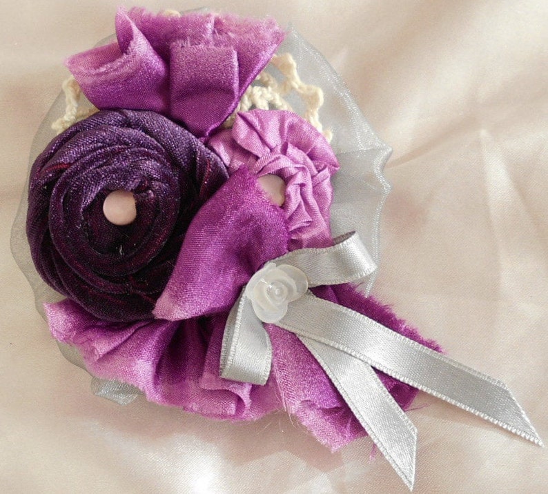 May include: A purple and silver fabric flower brooch with a white center. The flower is made of multiple layers of fabric and has a silver ribbon tied around it.