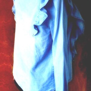 May include: Light blue long-sleeve shirt with ruffled detailing around the collar and shoulders. The shirt is made of a soft, lightweight fabric. The back of the shirt is visible, showing the collar and the ruffled design. The sleeves are long and cuffed.