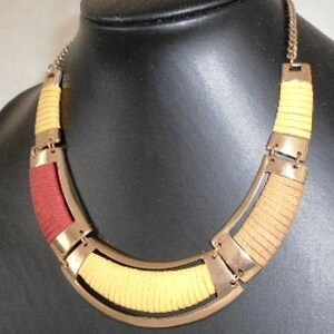 Puede incluir: Un collar de color dorado con un diseño geométrico. El collar está hecho de tiras superpuestas de tela marrón y amarilla.