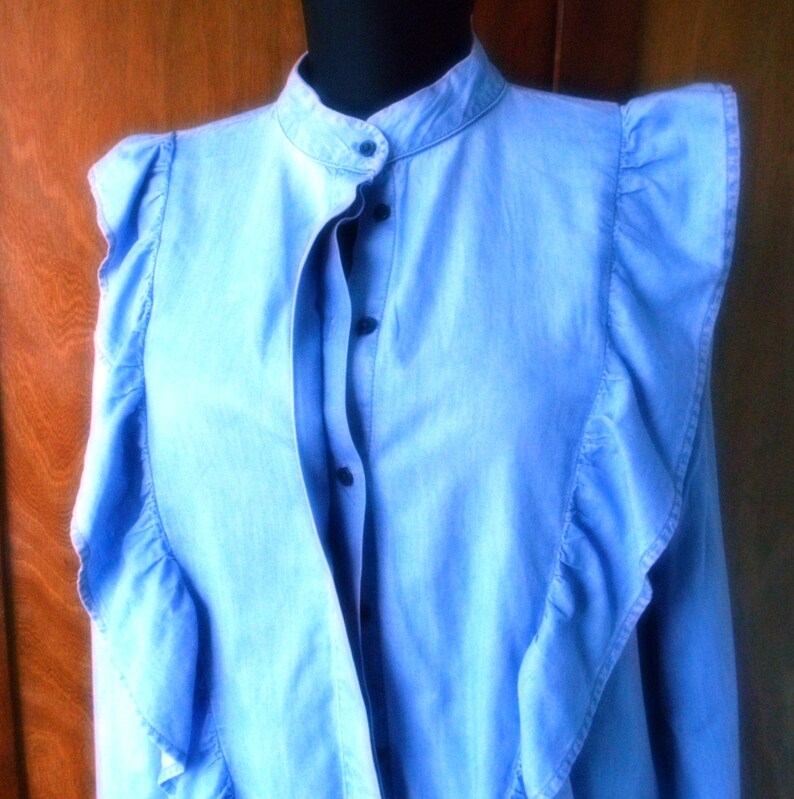 May include: Light blue denim shirt with a stand-up collar and ruffled accents. The shirt features a button-down front and long sleeves. The ruffles cascade down the front and around the shoulders, adding a decorative touch to the garment.