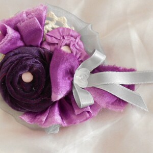 May include: A purple and silver fabric flower brooch with a white pearl center. The flower is made of multiple layers of fabric and is accented with a silver ribbon.