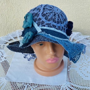 Puede incluir: Un sombrero azul para el sol con ala ancha y un acento floral decorativo. El sombrero presenta una corona de encaje y detalles de mezclilla, con un ala azul oscuro y una cinta azul claro. El sombrero se exhibe en una cabeza de maniquí.