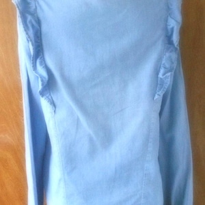 May include: Light blue denim shirt with long sleeves and ruffled shoulder detailing. The shirt is button-down with a classic collar and a curved hemline. The garment is displayed on a mannequin.
