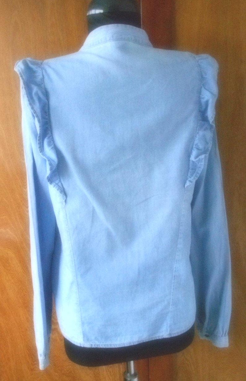 May include: Light blue denim shirt with long sleeves and ruffled shoulder detailing. The shirt is button-down with a classic collar and a curved hemline. The garment is displayed on a mannequin.