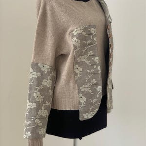 Peut inclure: Un pull en maille beige avec un panneau en tissu &agrave; motif floral sur le devant et les manches. Le pull a un col rond et des manches longues.