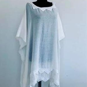 Op de afbeelding: Een witte, halfdoorzichtige poncho met een los, vloeiend silhouet. Het kledingstuk heeft een wijde halslijn en een onderzoom die beide zijn versierd met ingewikkelde witte gehaakte kanten randen in een geschulpt patroon. De stof is licht.