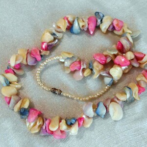 Puede incluir: Un collar de conchas multicolores con conchas rosas, blancas y azules. El collar est&aacute; hecho de conchas de forma irregular y est&aacute; enhebrado en un hilo delgado.