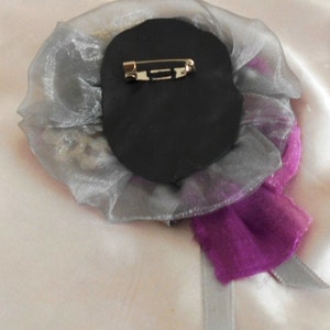 May include: A silver and purple fabric flower brooch with a black backing and a gold pin.