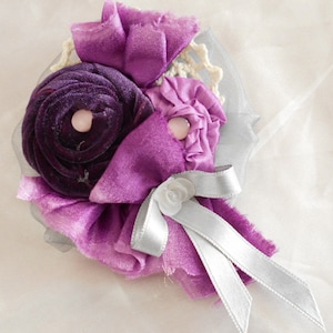 May include: A purple and silver fabric flower brooch with a white lace center. The brooch is made of multiple layers of fabric and has a satin ribbon.