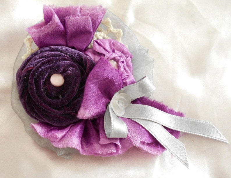 May include: A purple and silver fabric flower brooch with a velvet center. The flower is made of layers of fabric and has a delicate, feminine look.