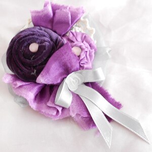 May include: A purple and white fabric flower brooch with a silver ribbon. The flower is made of layers of fabric and has a pearl-like center.