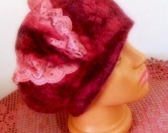 May include: A deep red felted beret featuring a textured surface and a decorative light pink lace trim attached to the side. The hat is displayed on a smooth, neutral-toned mannequin head, showcasing the soft, rounded shape of the felted material.