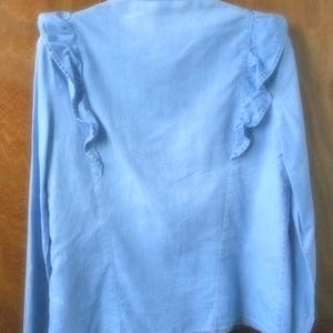 May include: Light blue denim shirt with long sleeves and ruffled shoulder detailing. The back of the shirt is visible, showcasing the ruffled accents. The shirt has a classic collar and button-down design, suitable for casual wear.