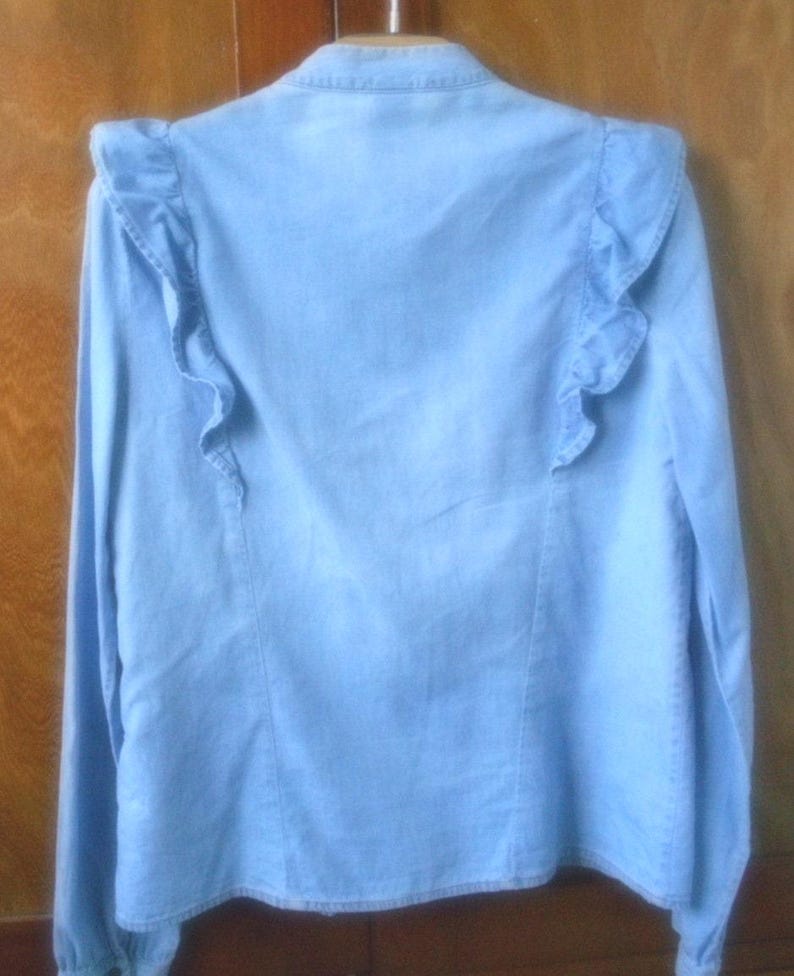 May include: Light blue denim shirt with long sleeves and ruffled shoulder detailing. The back of the shirt is visible, showcasing the ruffled accents. The shirt has a classic collar and button-down design, suitable for casual wear.