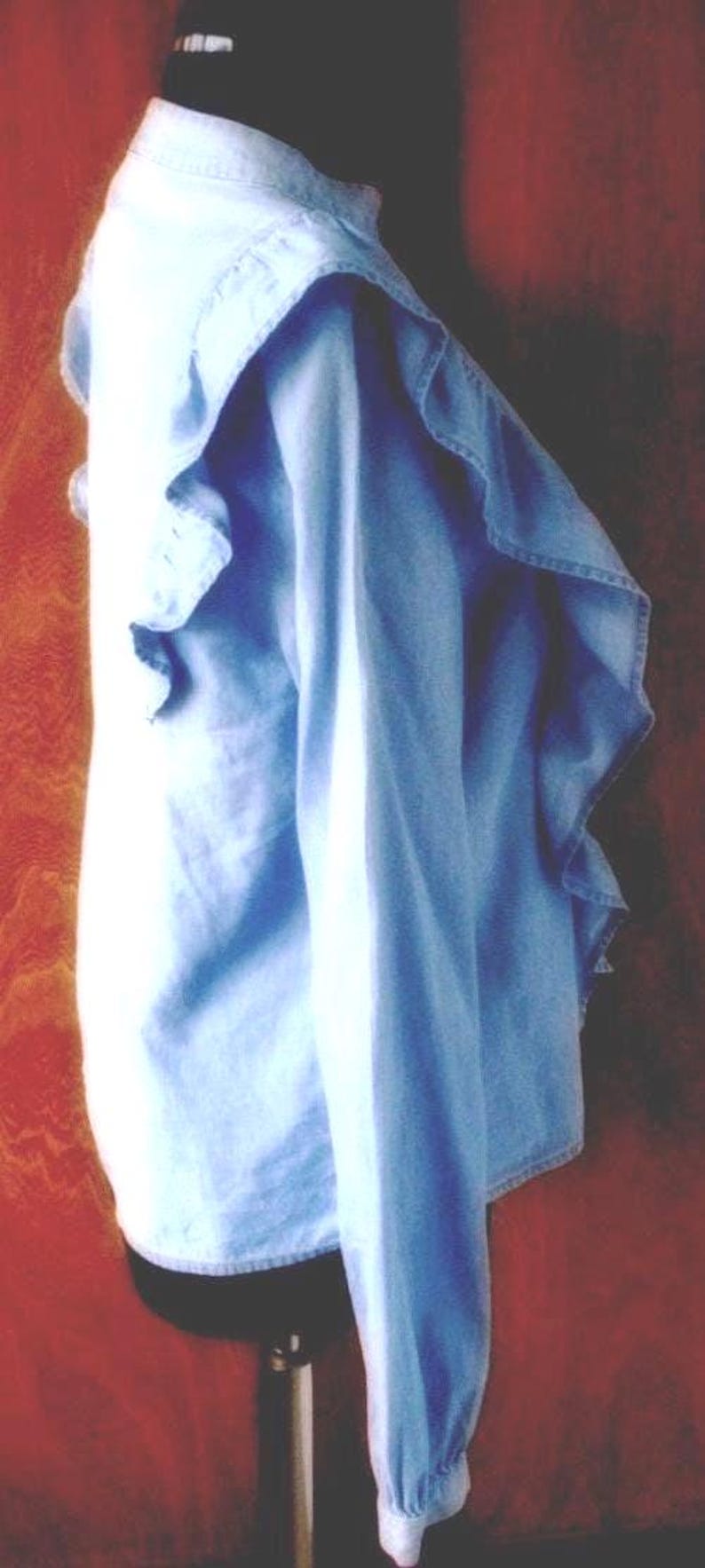 May include: Light blue denim shirt with ruffled detailing on the collar and sleeves. The shirt is displayed on a black mannequin. The background is a reddish-brown color.