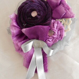 May include: A purple and silver fabric flower brooch with a silver ribbon bow. The flower is made of velvet and satin.