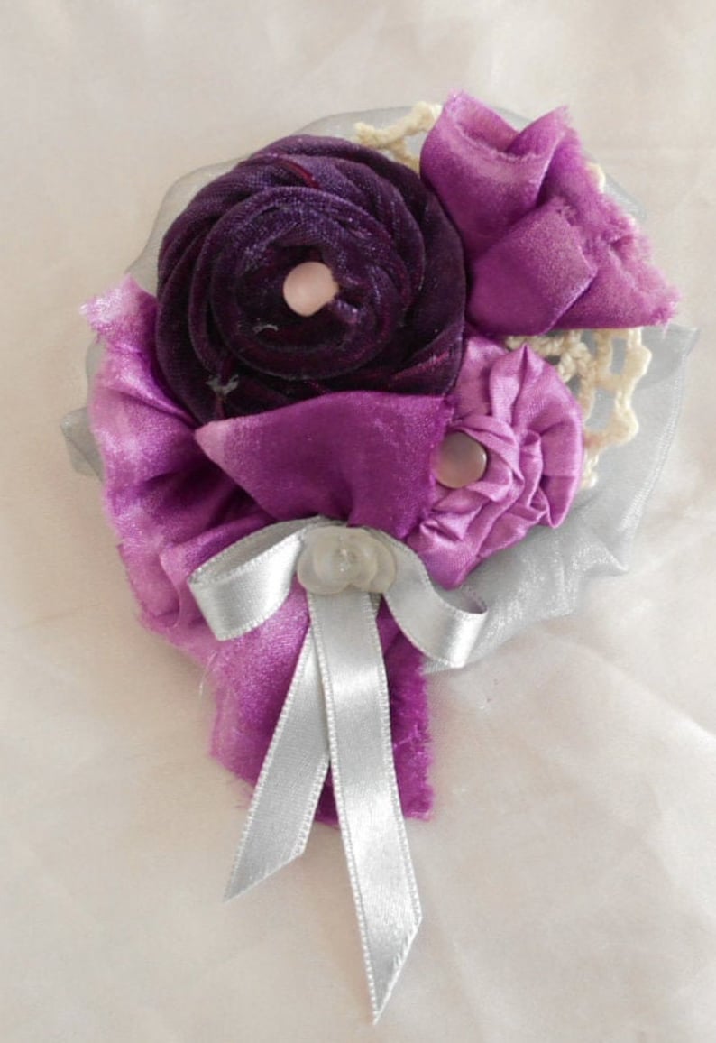 May include: A purple and silver fabric flower brooch with a silver ribbon bow. The flower is made of velvet and satin.