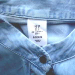 May include: Close-up of a light blue denim shirt collar with a button closure. A white tag displays size information: EUR 38, US 6, CA 6, CN 165/88A, MX 6, and the brand &DENIM. The shirt is made in India.