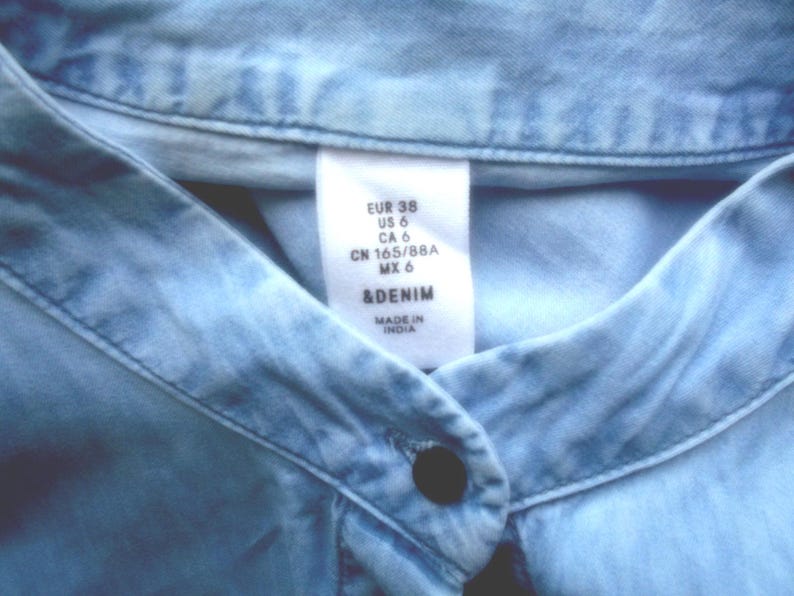 May include: Close-up of a light blue denim shirt collar with a button closure. A white tag displays size information: EUR 38, US 6, CA 6, CN 165/88A, MX 6, and the brand &DENIM. The shirt is made in India.