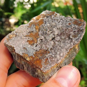 May include: A hand holds a piece of raw, natural stone. The stone has a rough, textured surface with a mix of colors, including brown, gray, and white. The top surface has a bubbly, crystal-like appearance. The edges of the stone are a darker brown.