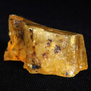 May include: A raw, translucent amber specimen with a golden-yellow hue, featuring irregular jagged edges and dark, natural inclusions embedded within the crystalline structure, showcasing a rough, unpolished surface texture throughout the piece.