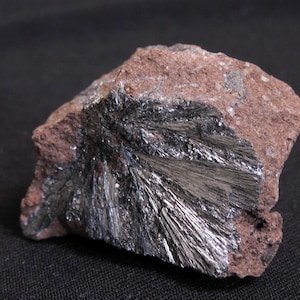 May include: A raw mineral specimen featuring a cluster of metallic, needle-like crystals in a dark silver hue embedded within a rough, porous, reddish-brown rock matrix. The crystalline formation radiates outward from a central point.