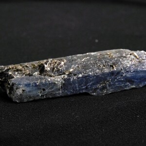 May include: A raw, elongated kyanite crystal specimen featuring a deep blue color gradient transitioning to translucent white at one end, with visible dark, metallic-looking mineral inclusions embedded along the textured, blade-like surface.