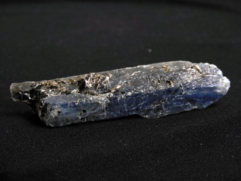 May include: A raw, elongated kyanite crystal specimen featuring a deep blue color gradient transitioning to translucent white at one end, with visible dark, metallic-looking mineral inclusions embedded along the textured, blade-like surface.