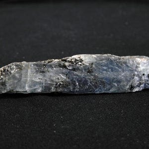 May include: A long, raw kyanite crystal specimen featuring a translucent blue and white color palette with visible dark mineral inclusions throughout its elongated, blade-like structure. The mineral displays a striated, layered texture.