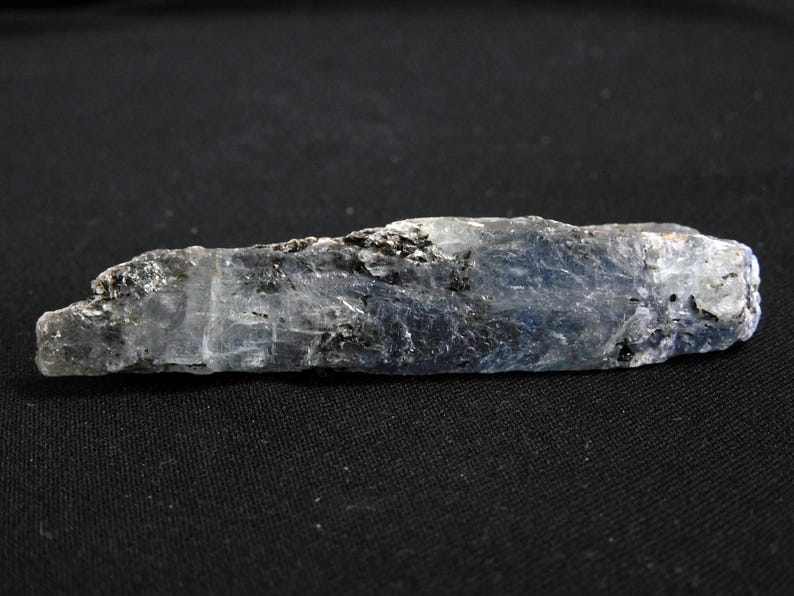 May include: A long, raw kyanite crystal specimen featuring a translucent blue and white color palette with visible dark mineral inclusions throughout its elongated, blade-like structure. The mineral displays a striated, layered texture.