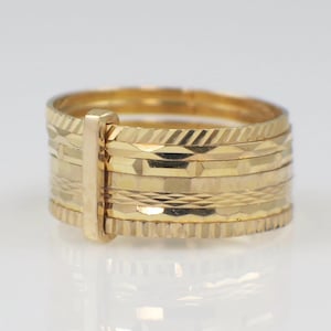 May include: A gold ring with multiple bands, each featuring a unique textured pattern. A vertical gold bar connects the bands, creating a cohesive design. The ring's surface reflects light, highlighting its intricate details and golden hue.
