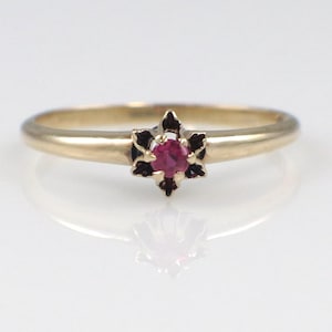 May include: A gold ring with a starburst design featuring a central red gemstone. The ring's band is a simple, polished gold, and the starburst setting has dark accents. The ring is displayed on a white surface, reflecting its shape.