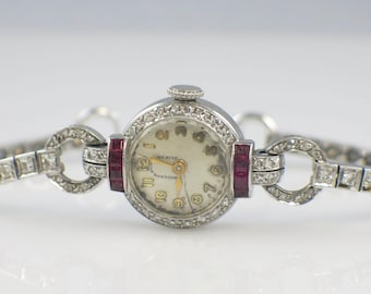 May include: A vintage silver wristwatch with a round face and a diamond-encrusted bezel. The watch features a cream-colored dial with gold hands and numerals. The band is also silver, with diamond accents and ruby-colored rectangular stones. The watch is a classic piece of jewelry.