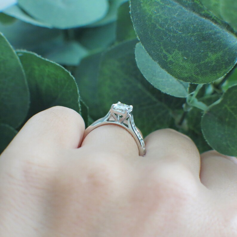May include: A silver engagement ring with a square-shaped diamond set in a delicate, intricate design. The ring is on a finger, with a green leafy background.