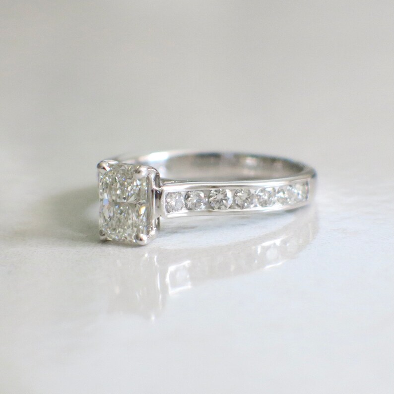 May include: A silver engagement ring with a large, clear, rectangular-shaped diamond set in a prong setting. The band is thin and has a row of smaller diamonds set in a channel setting.