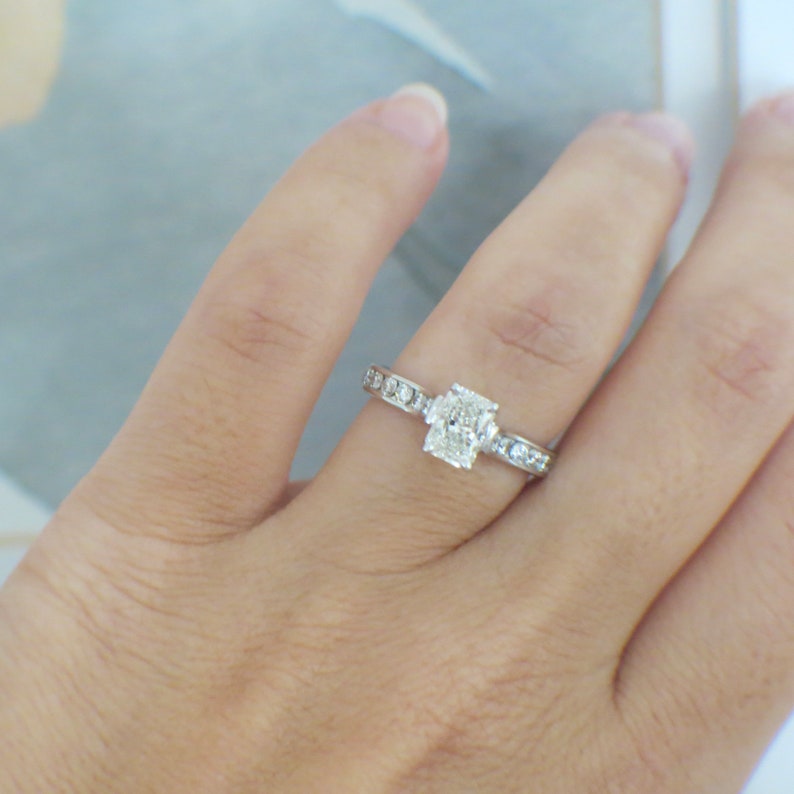 May include: A silver engagement ring with a large, rectangular-shaped diamond in the center and smaller diamonds set in a band.
