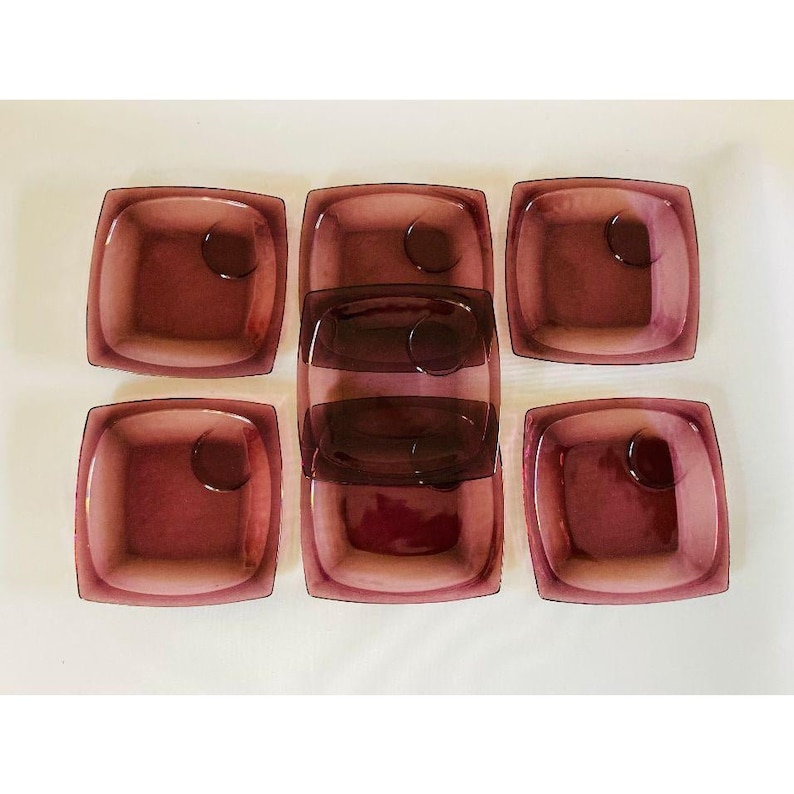 May include: Set of six square glass dishes in a deep purple hue. The dishes are stacked in pairs, with three pairs in a row. Each dish has a small circular indentation in the center.