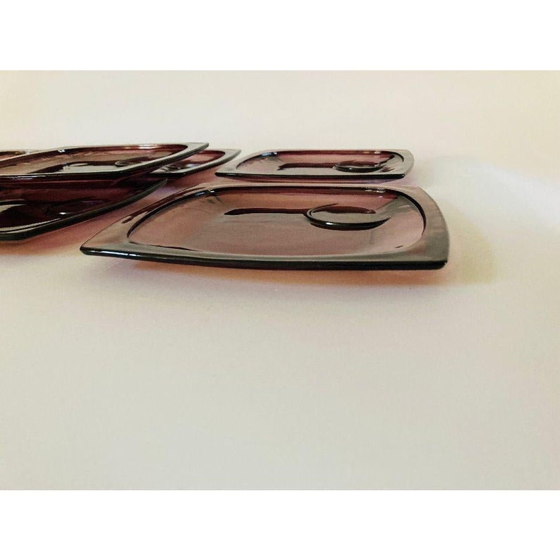 May include: Set of six purple glass plates with a square shape and a slight curve to the edges. The plates are stacked on top of each other.