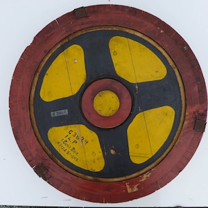 May include: A round wooden target with a red, black, and yellow design. The target has a center circle with a red ring around it. The target is labeled "Cone Box 4 Size Sticks C 33824 ILP".