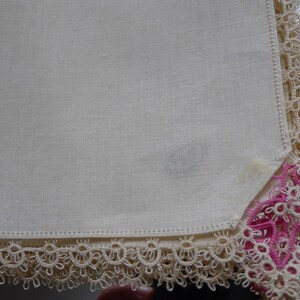 May include: A white linen handkerchief with a delicate lace border. The lace is cream colored with a pink floral design in the corner.