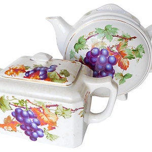 May include: Two white ceramic teapots with a grapevine and grape design. The teapots have a lid and a spout.