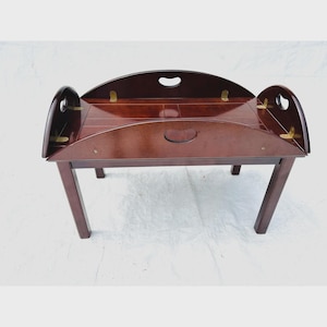 May include: A dark brown wooden butler tray table with a rectangular surface and four straight legs. The tray features high, curved sides with integrated handles and brass hinges connecting the folding panels to the main frame structure.