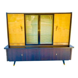 May include: A mid-century modern cabinet with a dark wood finish and sliding glass doors. The cabinet has a yellow wood veneer on the sides and a lower section with three drawers.