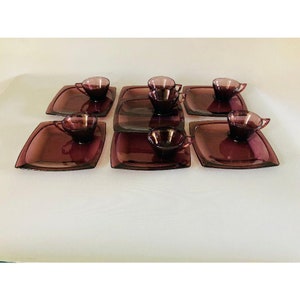 May include: Set of six purple glass teacups and saucers. Each saucer has a square shape with rounded corners. The cups are small and have a rounded shape.