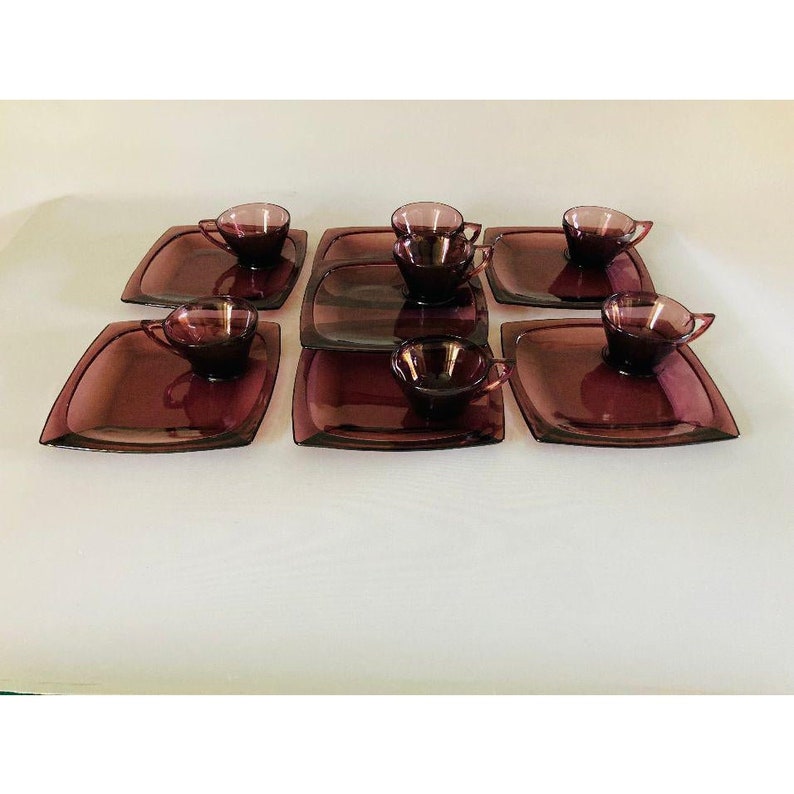 May include: Set of six purple glass teacups and saucers. Each saucer has a square shape with rounded corners. The cups are small and have a rounded shape.