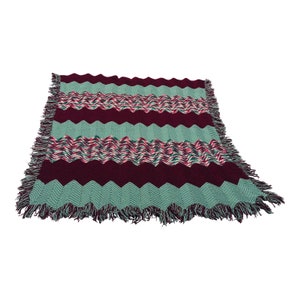 May include: A crocheted throw blanket with a chevron pattern in shades of burgundy, teal, and white. The blanket has a fringe border.