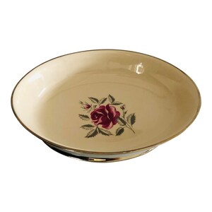 May include: Cream-colored oval serving dish with a gold rim and a floral rose design. The rose is a deep burgundy color, surrounded by green leaves. The dish has a slightly raised base and is likely made of ceramic or porcelain.